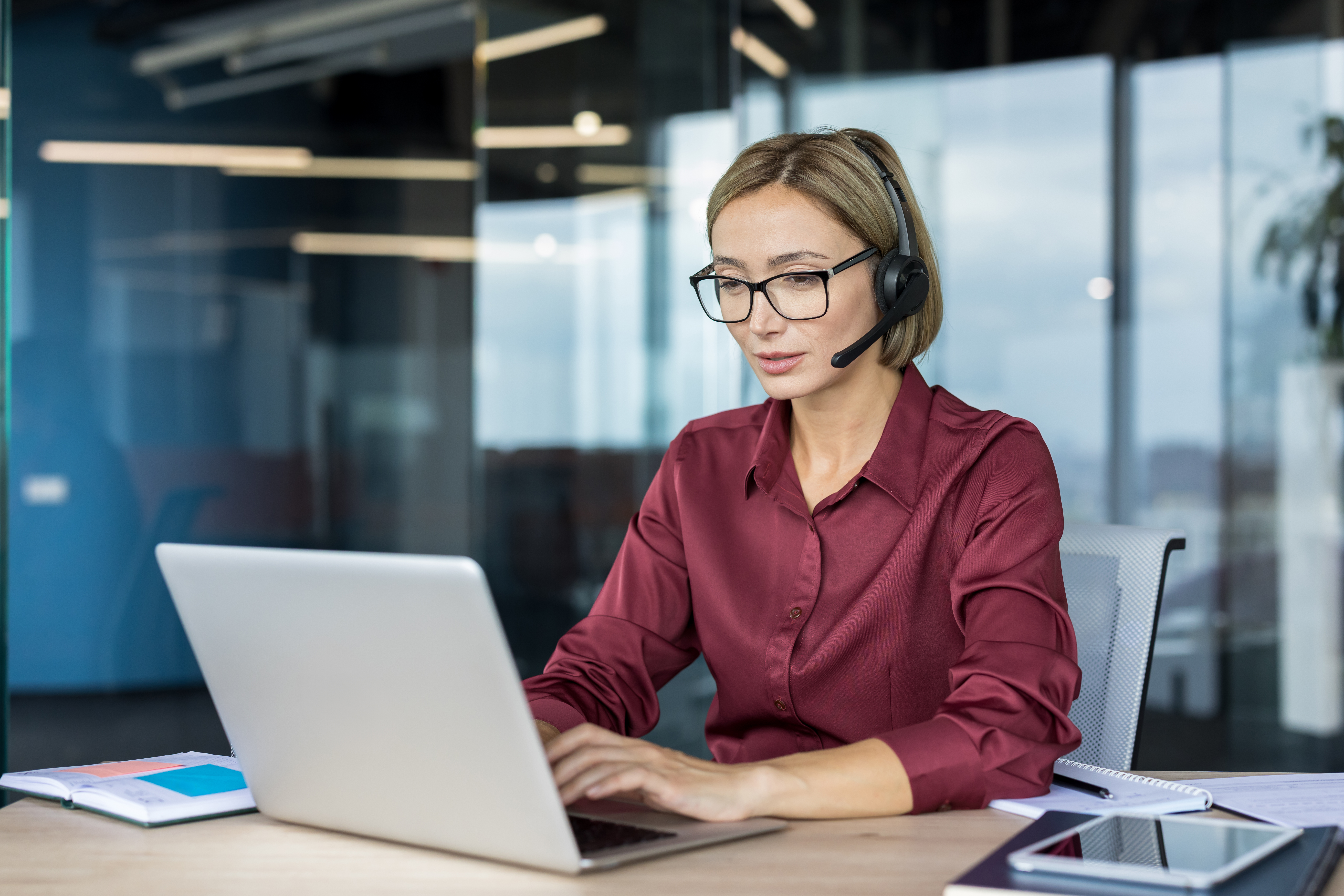 Young woman working as a customer service representative or support specialist, wearing a headset and spectacles while typing on a laptop computer at a desk in a contemporary office environment